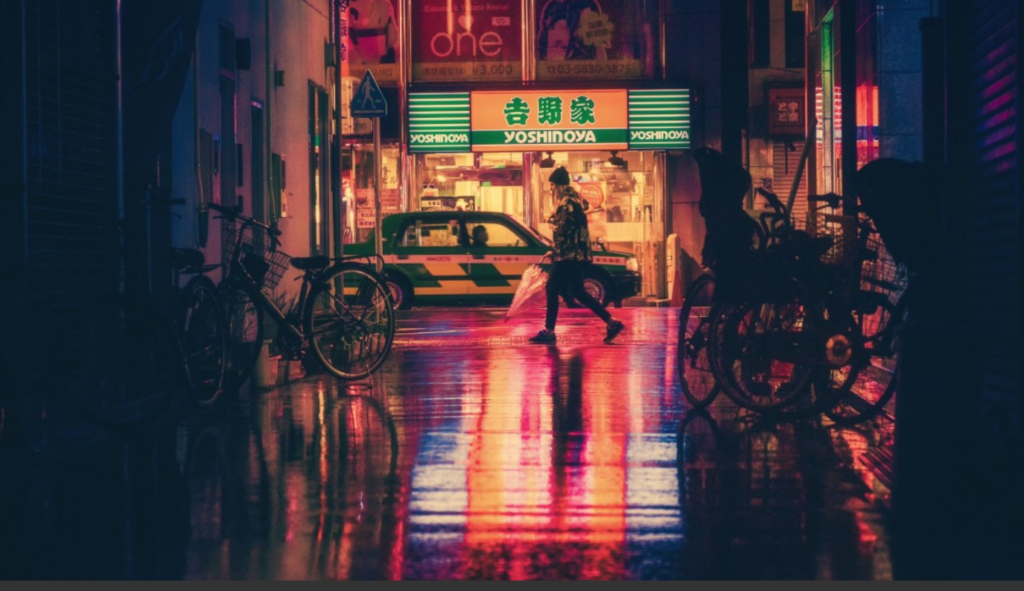 A person walking with an umbrella on a wet street reflecting neon lights, with bicycles parked nearby and a shop in the background.