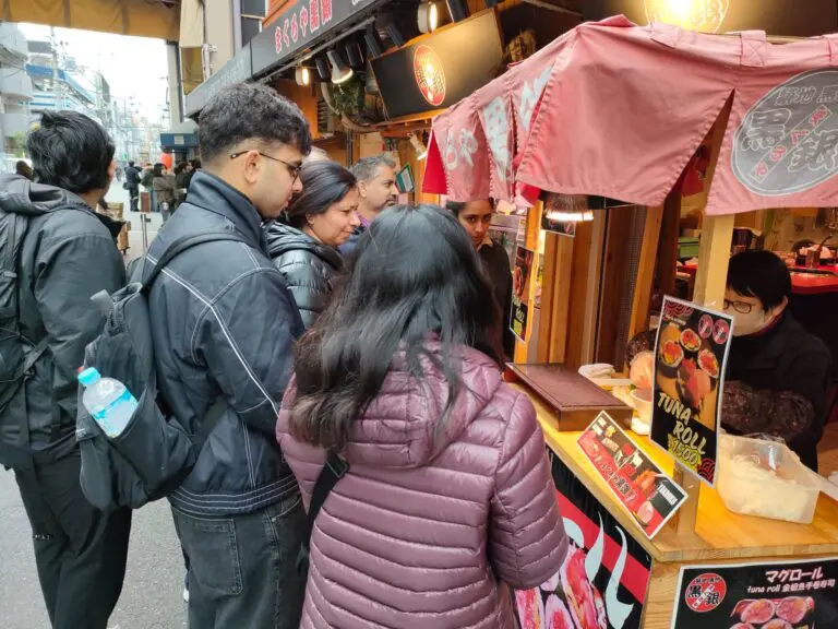 Group of people gathered around a food stall in a market, observing menu items and interacting with the vendor.