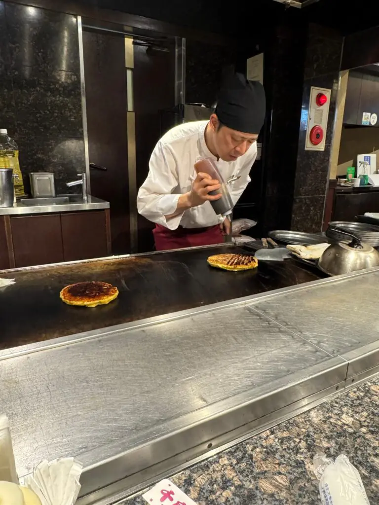 Chef preparing okonomiyaki on a teppanyaki grill with two cooked pancakes visible on the surface.