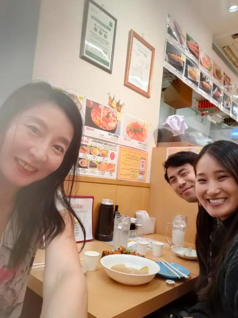 Three individuals smiling together at a dining table in a restaurant, with menus and food items visible in the background.