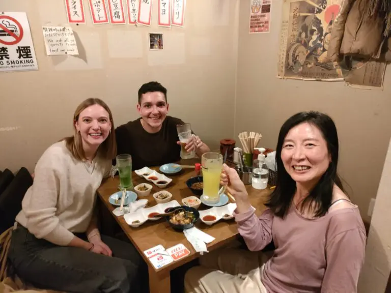 Three individuals sitting at a table in a restaurant, each holding a drink, with various dishes and condiments on the table.