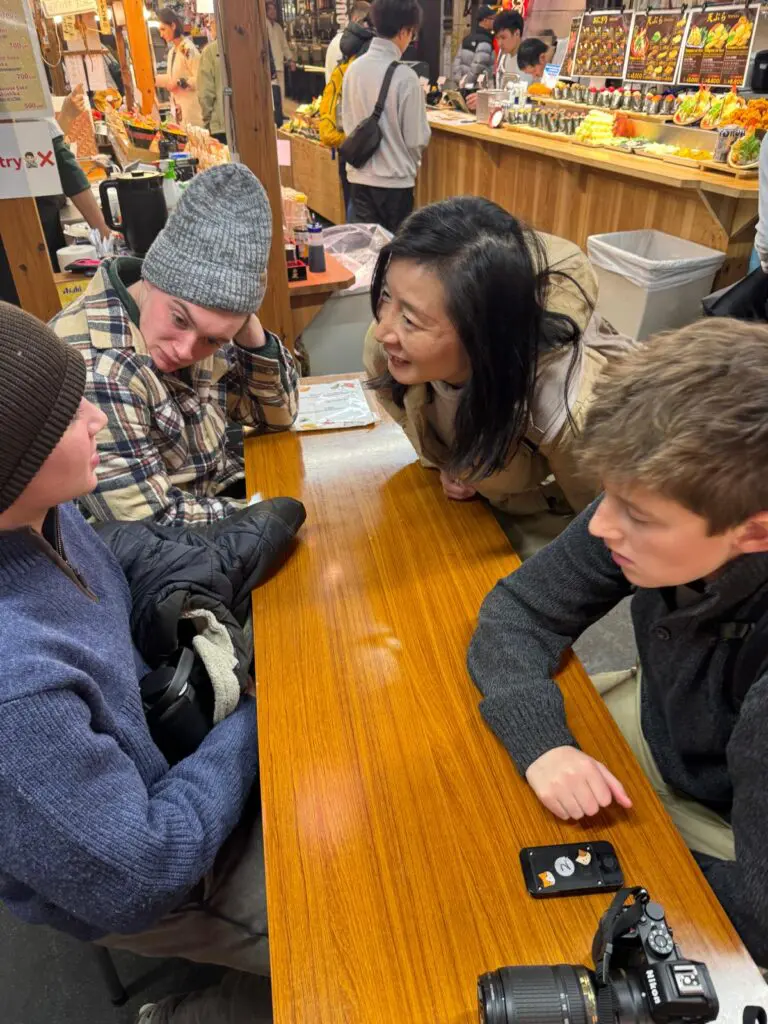 Group of four individuals engaged in conversation at a wooden table in a market setting.