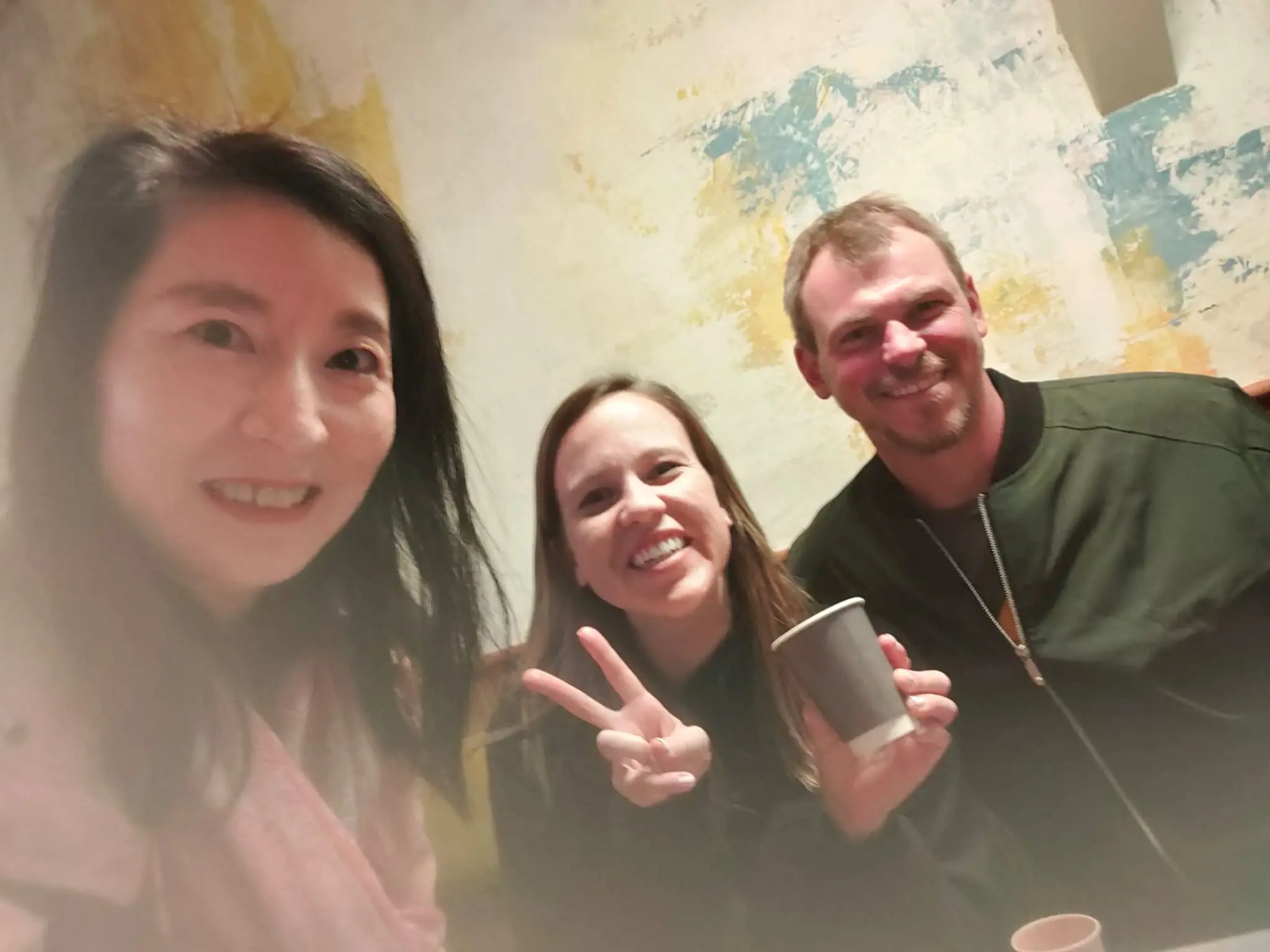 Three individuals posing for a photo, with one holding a cup and making a peace sign, in a softly lit setting.