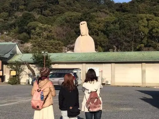 Walking with the Kyoto Hidden Gems Tour group toward a white Ryozan Kannon statue, framed by green forest hills under a clear blue sky in Kyoto today.