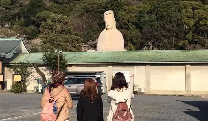 Walking with the Kyoto Hidden Gems Tour group toward a white Ryozan Kannon statue, framed by green forest hills under a clear blue sky in Kyoto today.
