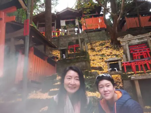 Jane and another individual posing at Fushimi Inari Shrine, surrounded by vibrant red torii gates and autumn leaves.