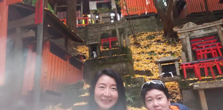 Jane and another individual posing at Fushimi Inari Shrine, surrounded by vibrant red torii gates and autumn leaves.