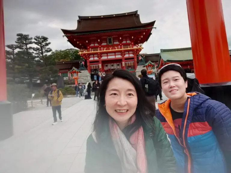 Jane and another individual posing at the entrance of Fushimi Inari Shrine, featuring traditional Japanese architecture and vibrant red torii gates.