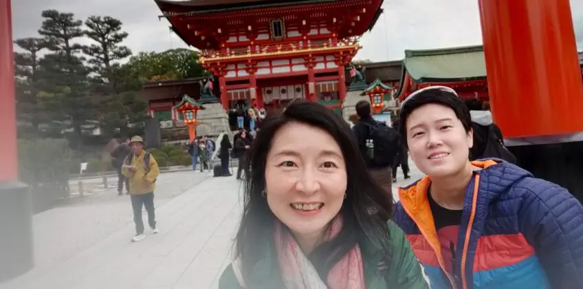 Jane and another individual posing at the entrance of Fushimi Inari Shrine, featuring traditional Japanese architecture and vibrant red torii gates.