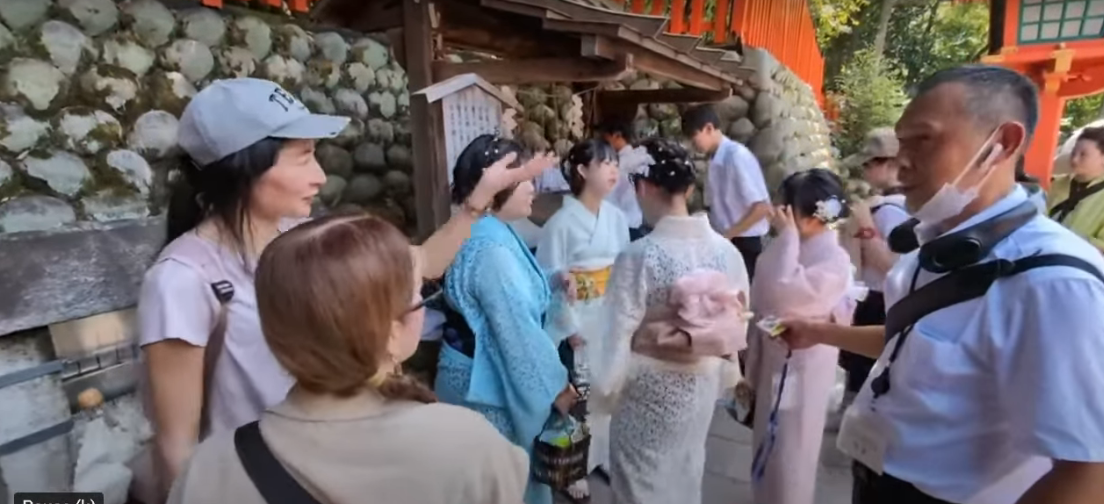 Group of individuals in traditional Japanese attire at Fushimi Inari Shrine, with one person gesturing while others engage in conversation.