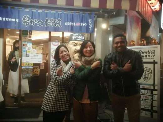 Three individuals posing in front of a restaurant with a sign in Japanese, showcasing a friendly interaction.