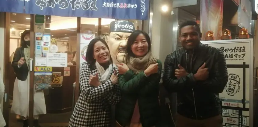 Three individuals posing in front of a restaurant with a sign in Japanese, showcasing a friendly interaction.