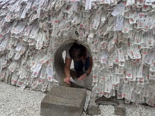 Kyoto’s Yasui Konpiragu stone prayer well packed with white ema plaques with red seals and calligraphy, with green foliage behind the shrine