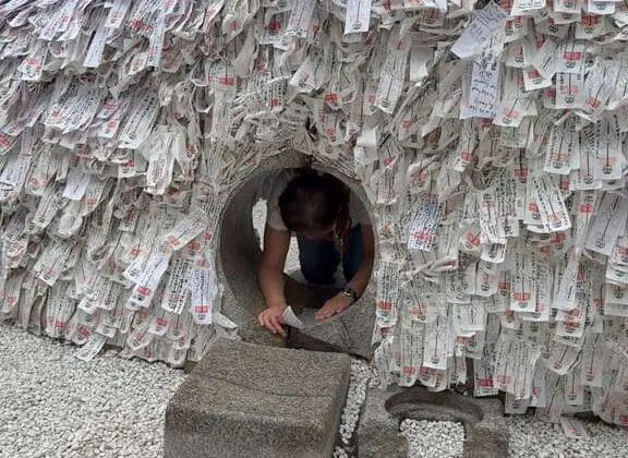 Kyoto’s Yasui Konpiragu stone prayer well packed with white ema plaques with red seals and calligraphy, with green foliage behind the shrine