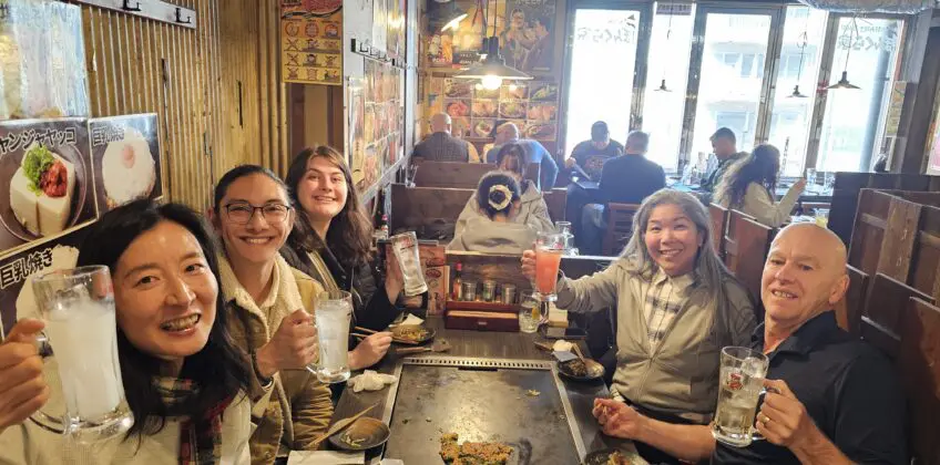 Alene Alaklawa and friends raise glasses at a teppanyaki table in Osaka, Japan, inside a wooden-walled restaurant decorated with posters and pendant lighting.Osaka Food Guide
