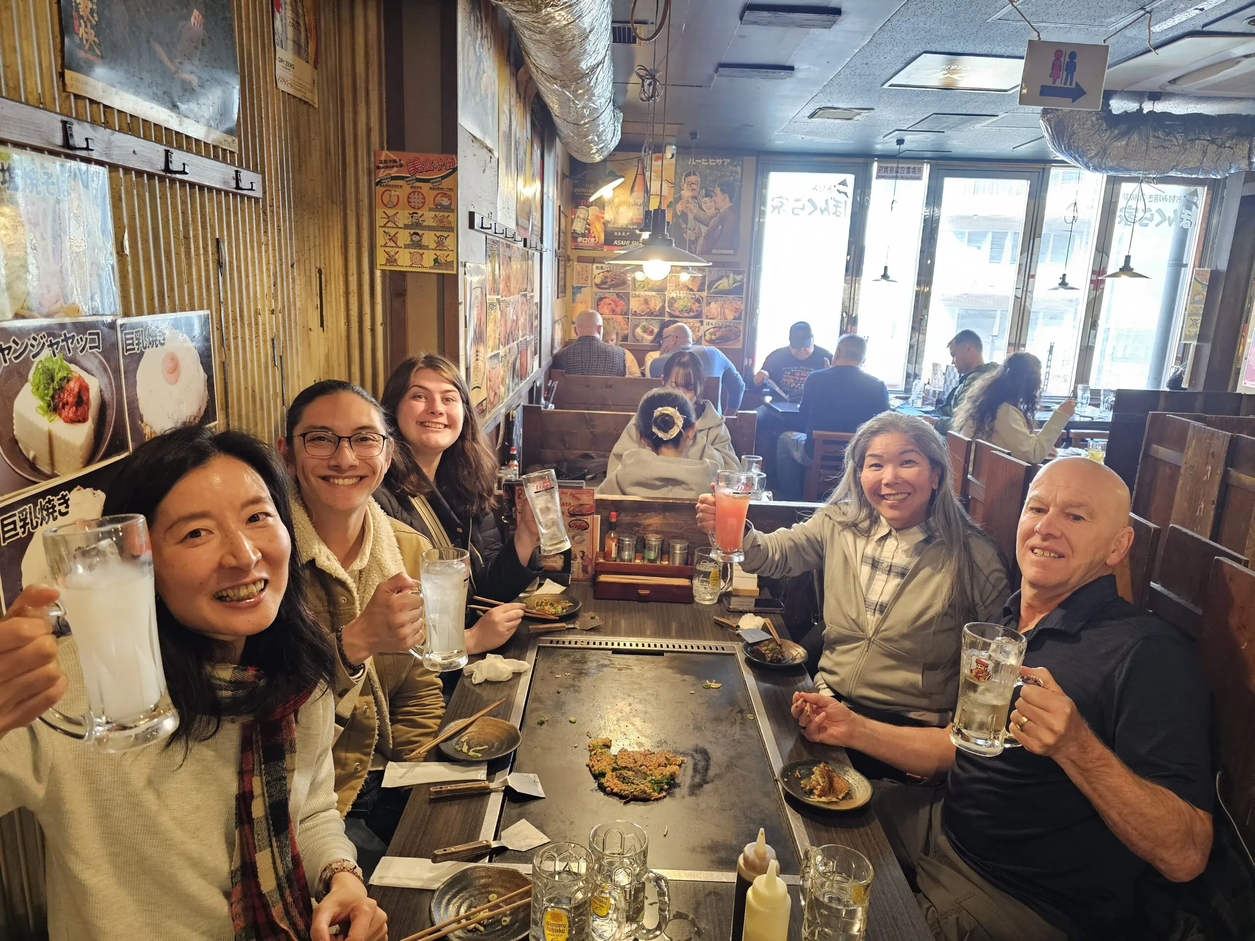 Alene Alaklawa and friends raise glasses at a teppanyaki table in Osaka, Japan, inside a wooden-walled restaurant decorated with posters and pendant lighting.Osaka Food Guide