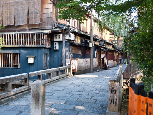 Kyoto street lined with traditional wooden machiya houses and a stone-paved path, framed by greenery in the historic Gion area of Kyoto, Japan.