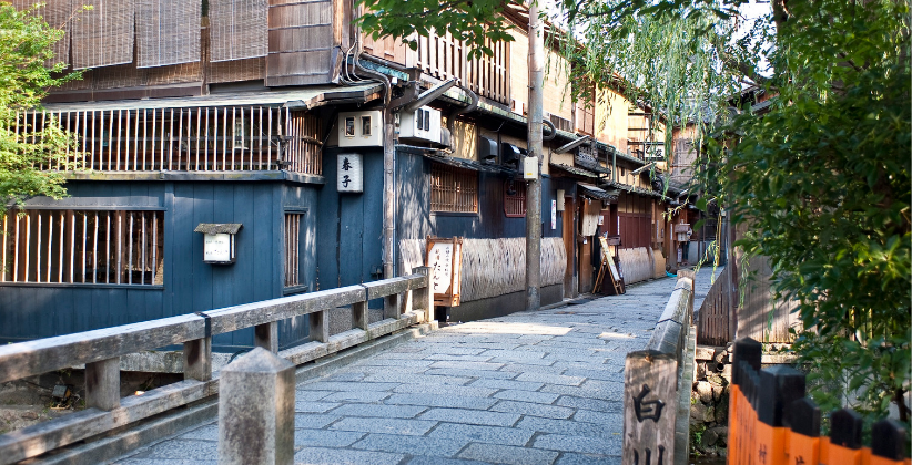 Kyoto street lined with traditional wooden machiya houses and a stone-paved path, framed by greenery in the historic Gion area of Kyoto, Japan.