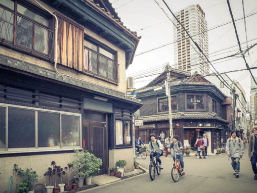 Urban street scene in Osaka with traditional wooden shopfronts, cyclists and pedestrians on a narrow road, overhead power lines, potted plants, and taller buildings in the background