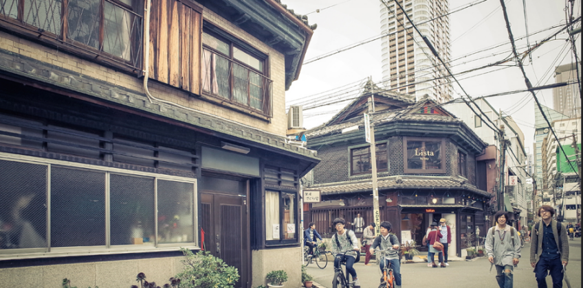 Urban street scene in Osaka with traditional wooden shopfronts, cyclists and pedestrians on a narrow road, overhead power lines, potted plants, and taller buildings in the background