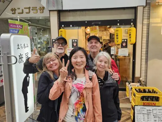 Kelly and a small group in Tenma, Osaka outside a shop entrance with Japanese signage and yellow lanterns, smiling and posing with peace signs