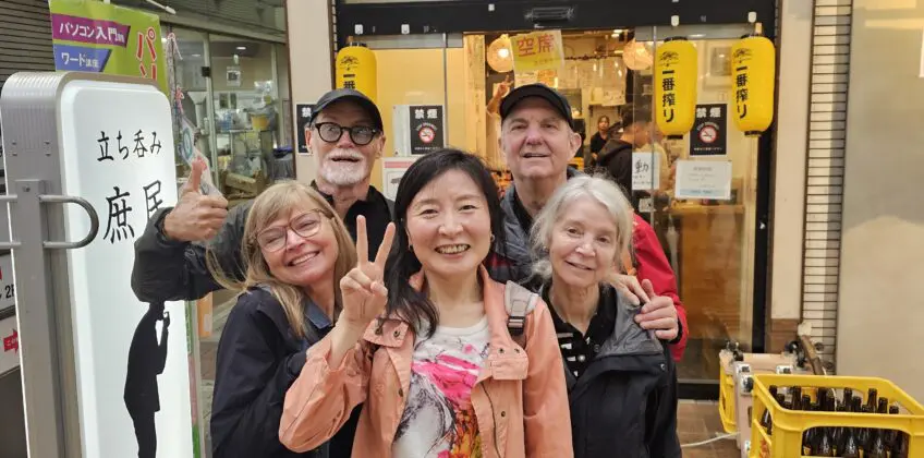 Kelly and a small group in Tenma, Osaka outside a shop entrance with Japanese signage and yellow lanterns, smiling and posing with peace signs