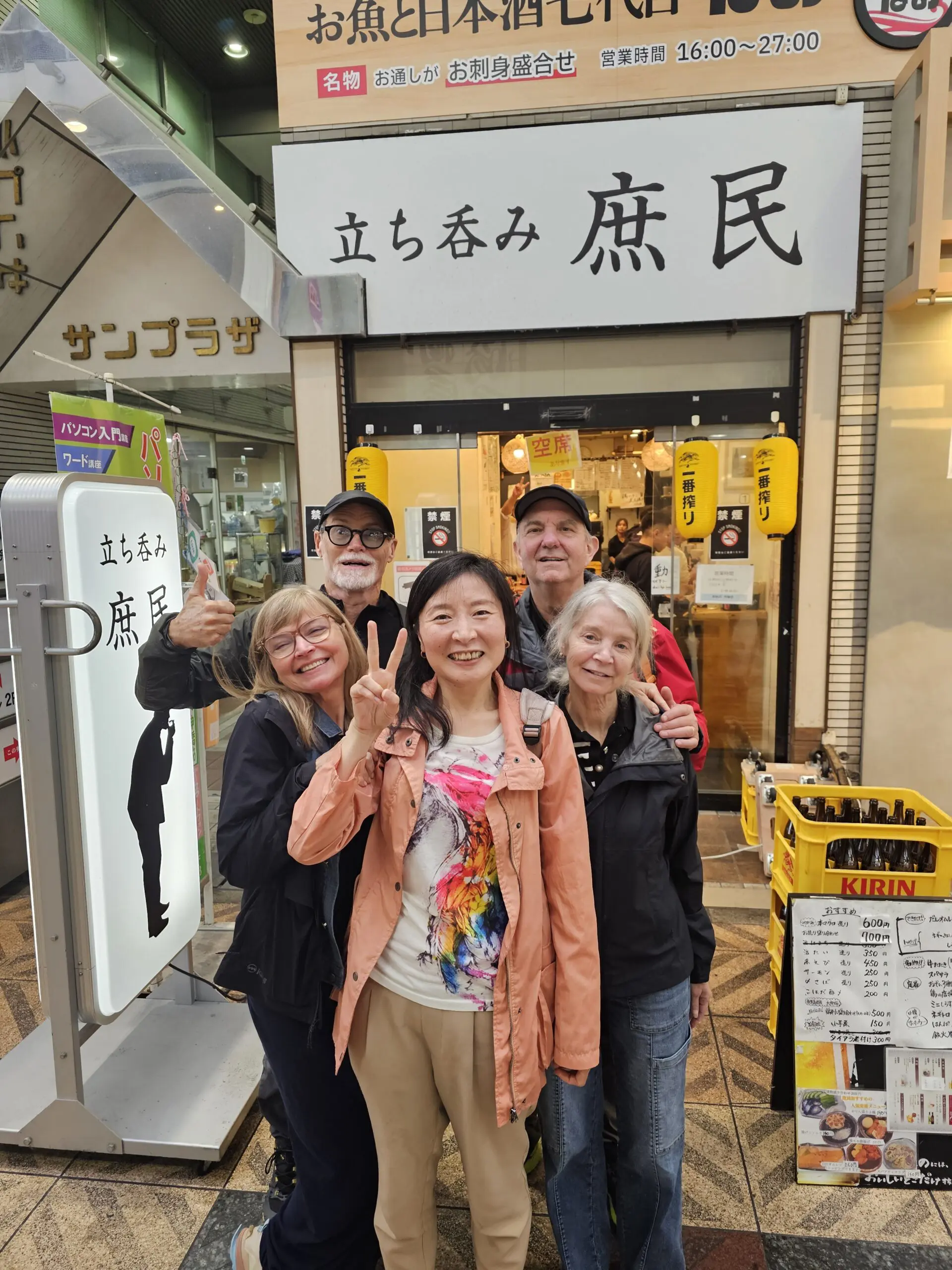 Kelly and a small group in Tenma, Osaka outside a shop entrance with Japanese signage and yellow lanterns, smiling and posing with peace signs