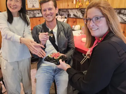 Courtney at Kuromon Market in Osaka inside a wood-paneled food stall with red signage, serving chopsticks and a seafood dish with friends.