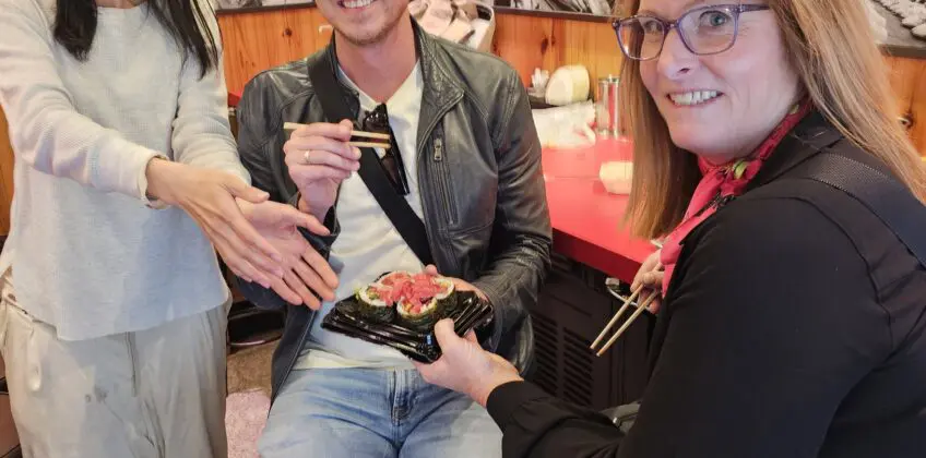 Courtney at Kuromon Market in Osaka inside a wood-paneled food stall with red signage, serving chopsticks and a seafood dish with friends.