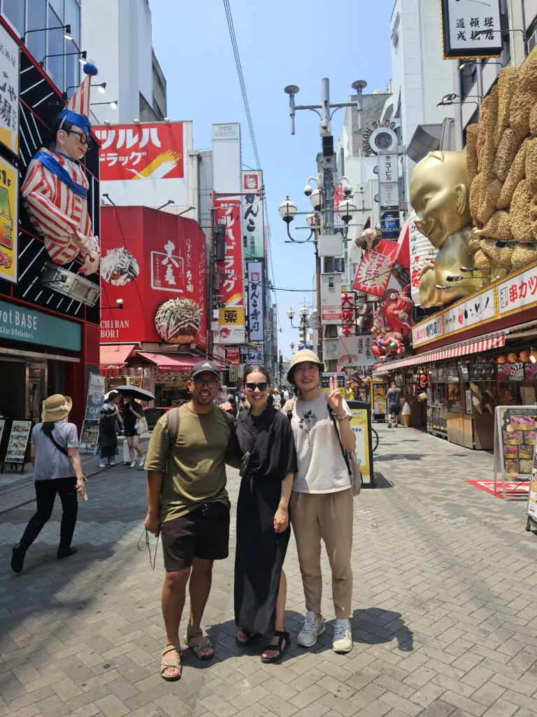 Three travelers pose on a lively Dotonbori street in Osaka, surrounded by bright signs, shops, and street food stalls.
