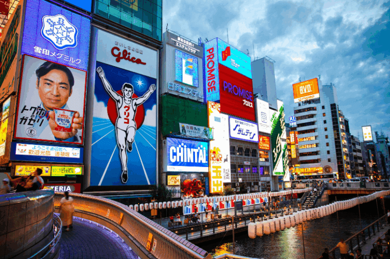 Osaka Tourist Attractions, Bright neon signs and advertisements illuminating a cityscape at dusk with a river in the foreground.