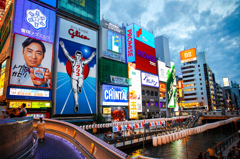Kansai Travel Blogs 4 Osaka Tourist Attractions, Bright neon signs and advertisements illuminating a cityscape at dusk with a river in the foreground.