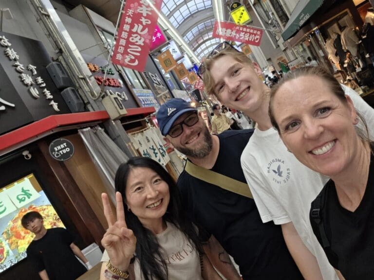 whatsapp-image-2025-06-29-at-04-52-08_cbbce02a Four individuals smiling together in a vibrant shopping district with colorful storefronts in the background.