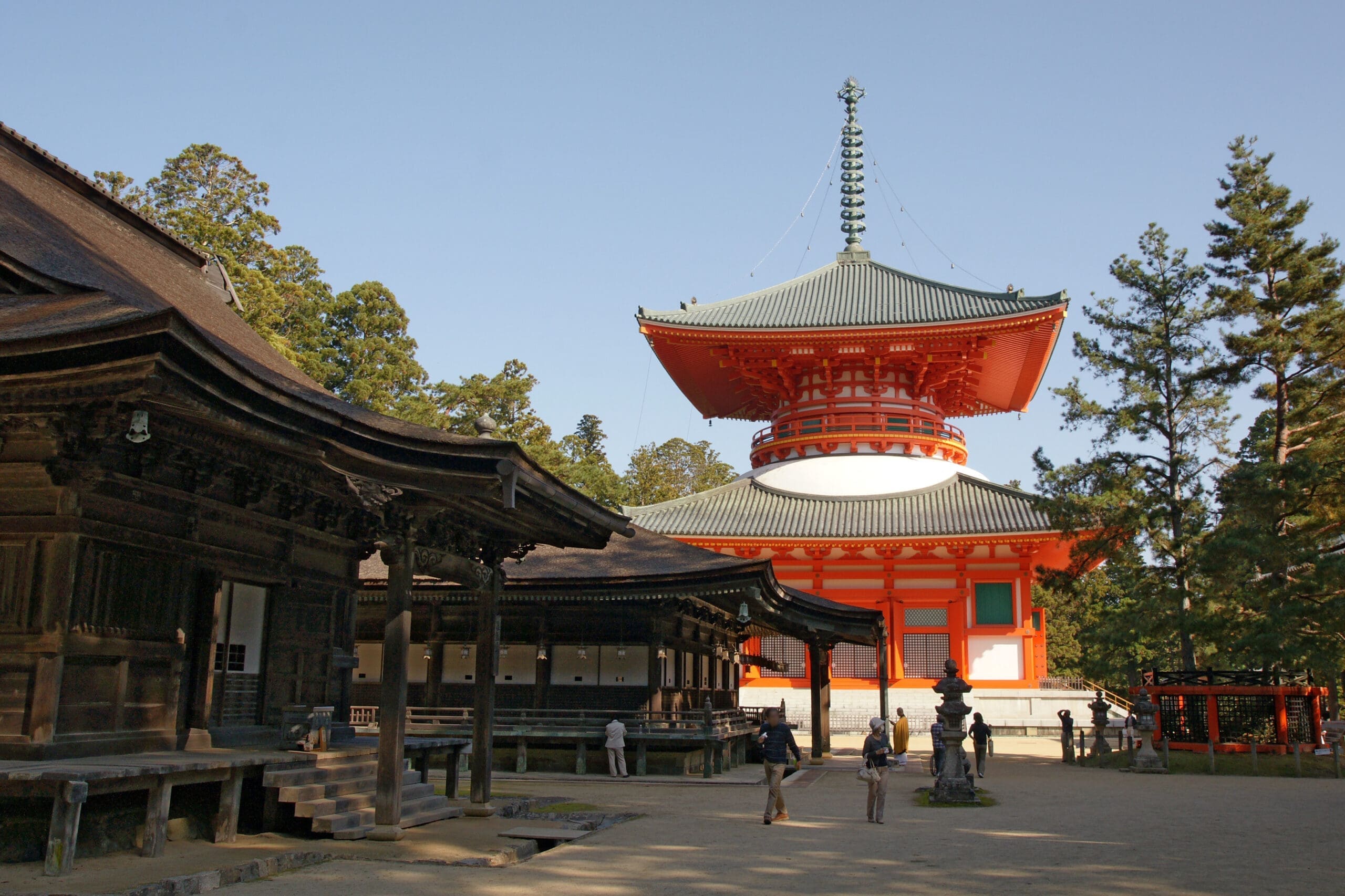 Pagoda structure with a green roof and red walls, surrounded by traditional wooden buildings in a serene outdoor setting.