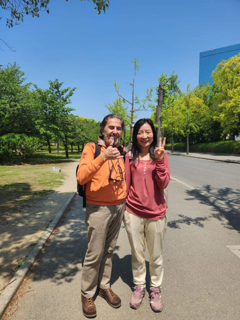whatsapp-image-2025-06-29-at-06-49-09_3291ebc6 Two individuals standing outdoors, one wearing an orange sweater and the other in a pink top, both smiling and posing for the camera.