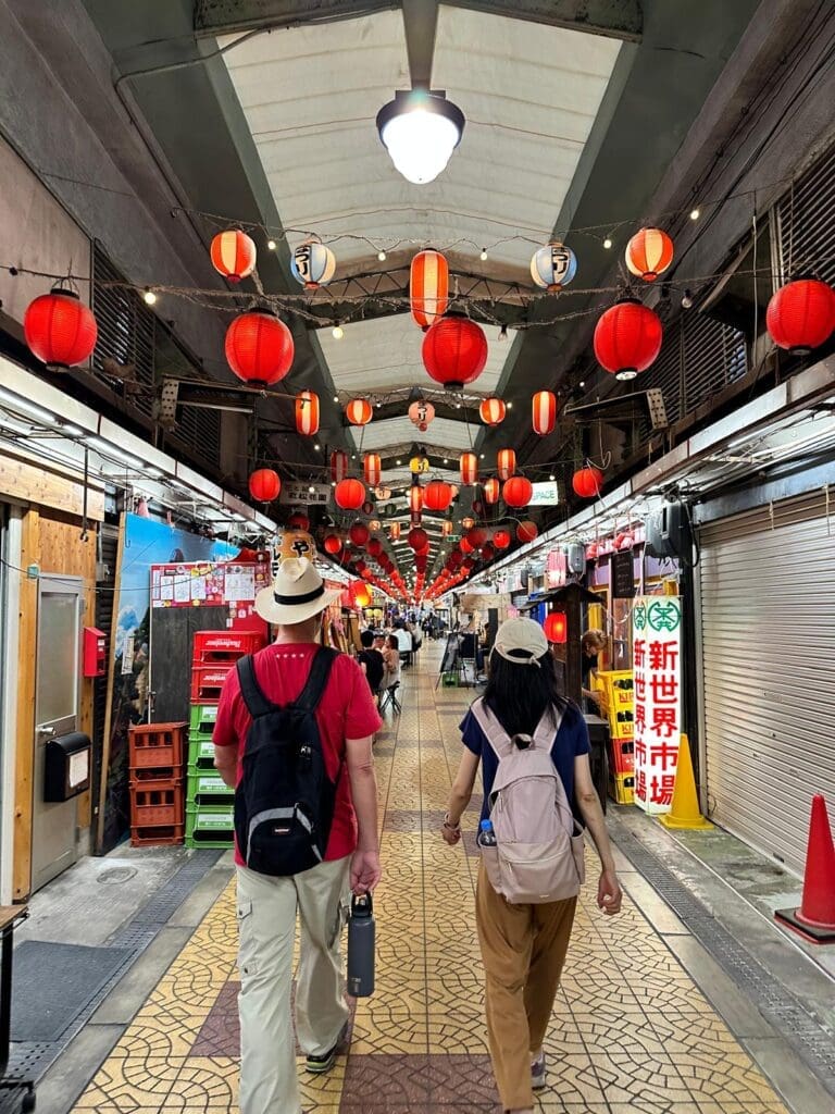 whatsapp-image-2025-06-29-at-06-49-11_e8522c50 Two individuals walking through a market street adorned with red lanterns and colorful storefronts.