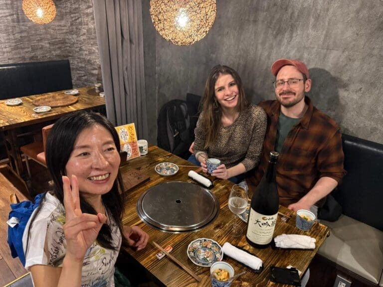 whatsapp-image-2025-07-15-at-10-26-52-am-1 Three individuals sitting at a table in a restaurant with food and drinks, one person making a peace sign.