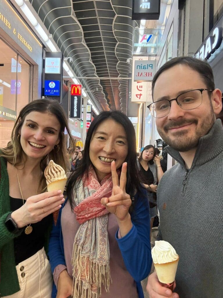 whatsapp-image-2025-07-15-at-10-26-52-am Three individuals holding ice cream cones in a shopping area with various store signs in the background.