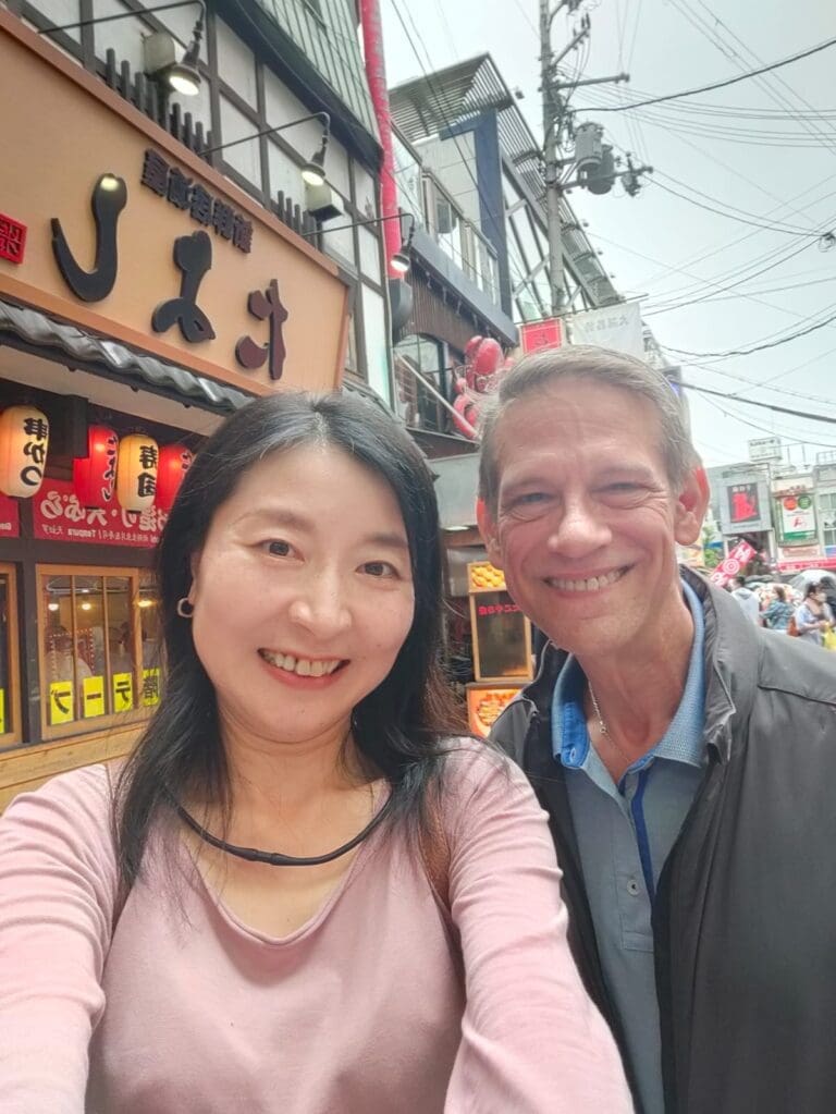 whatsapp-image-2025-07-15-at-10-26-54-am-2 Two individuals posing for a selfie in front of a restaurant with Japanese signage and a bustling street atmosphere.