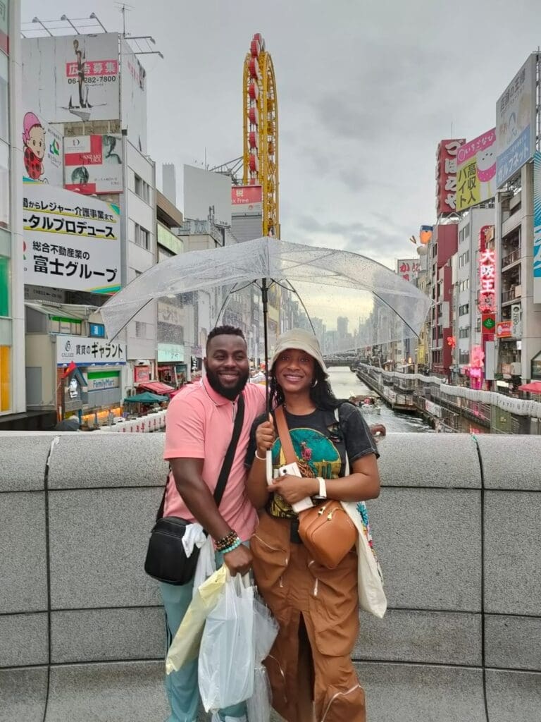 whatsapp-image-2025-07-15-at-10-26-54-am Two individuals holding an umbrella while standing on a bridge in a city with tall buildings and a canal.