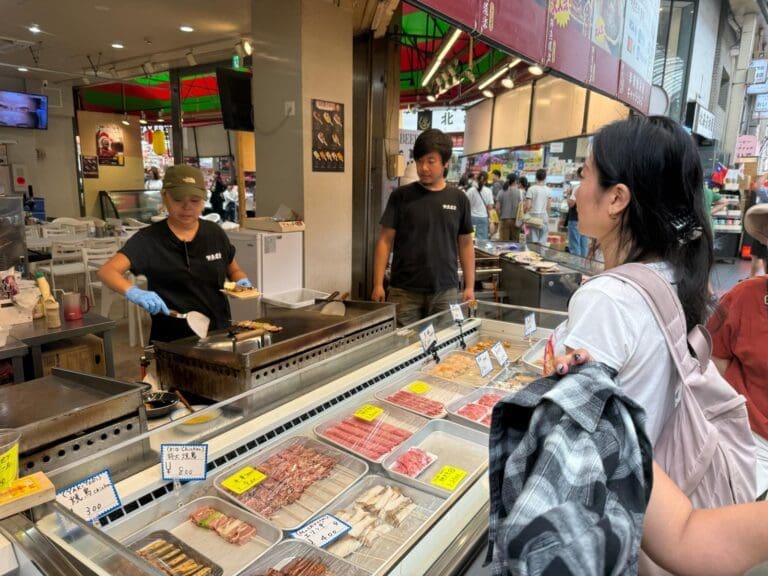 A vendor cooking food at a market stall while a customer observes, showcasing various food items displayed.