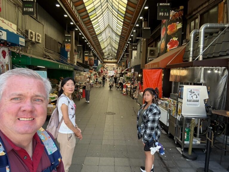 Two individuals standing in a bustling market with various food stalls and shops in the background.