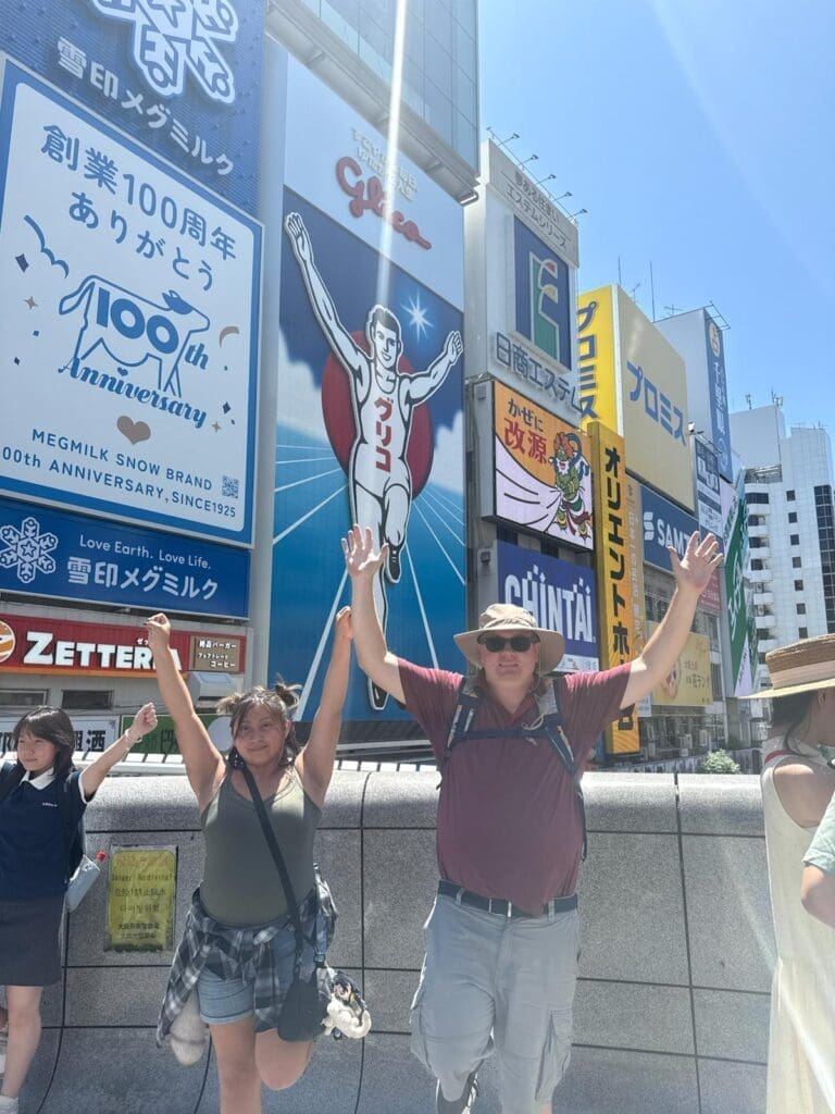 whatsapp-image-2025-07-15-at-10-27-09-am-2 Two individuals posing with arms raised in front of large advertisements in a city setting.