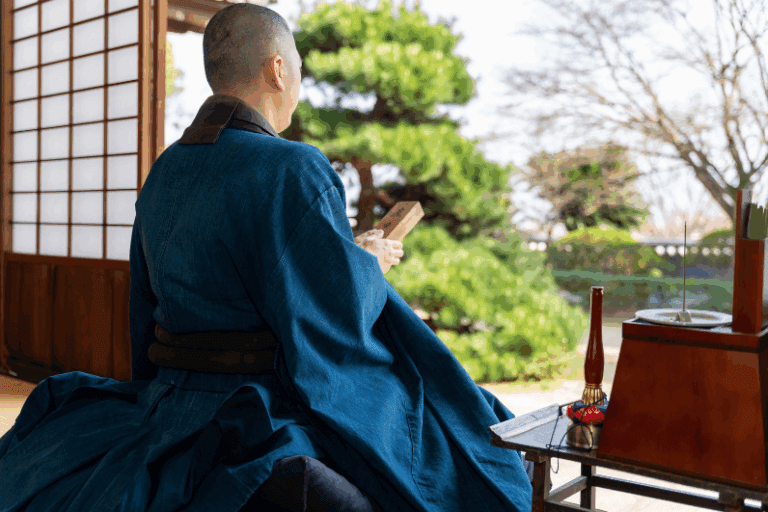 Koyasan from Osaka, A monk wearing traditional blue robes seated with a wooden object in hand, facing a garden view.