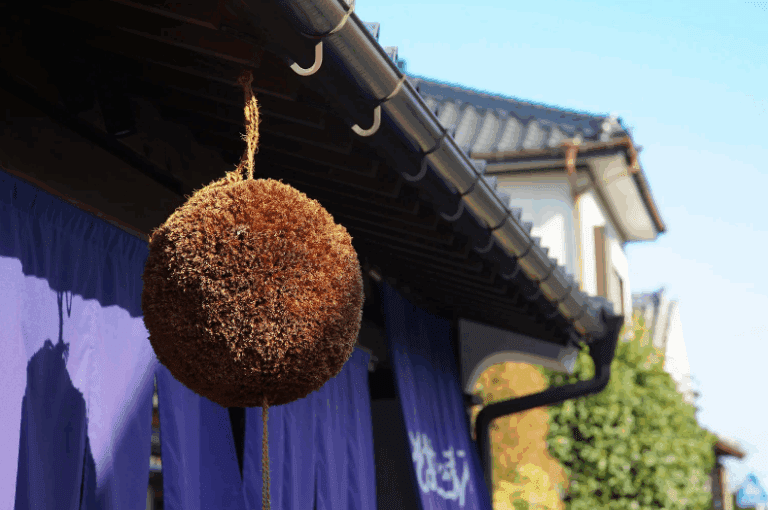 Kobe Sake Brewery, A round natural fiber ball hanging from a roof eave, with a blue sky in the background.