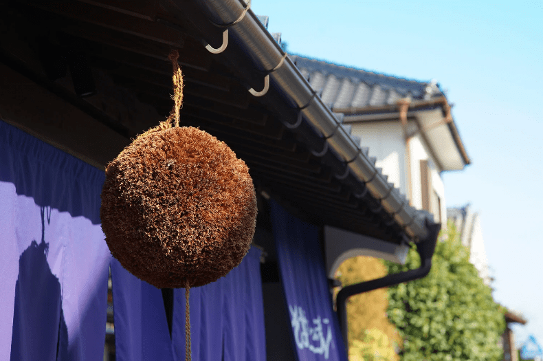 Kansai Travel Blogs 17 Kobe Sake Brewery, A round natural fiber ball hanging from a roof eave, with a blue sky in the background.
