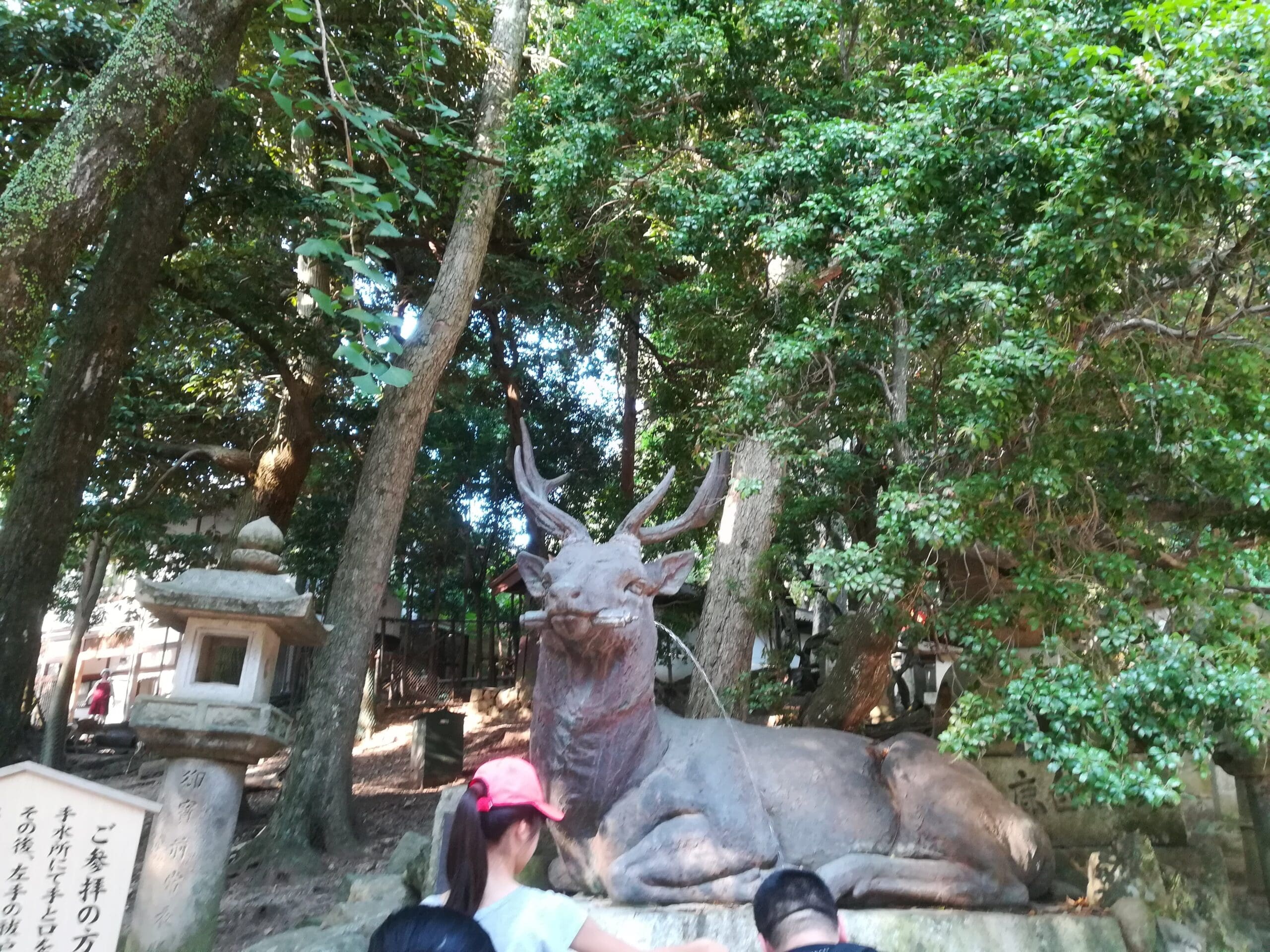 Bronze deer statue resting among trees at Kasuga Taisha in Nara, Japan, with visitors nearby.