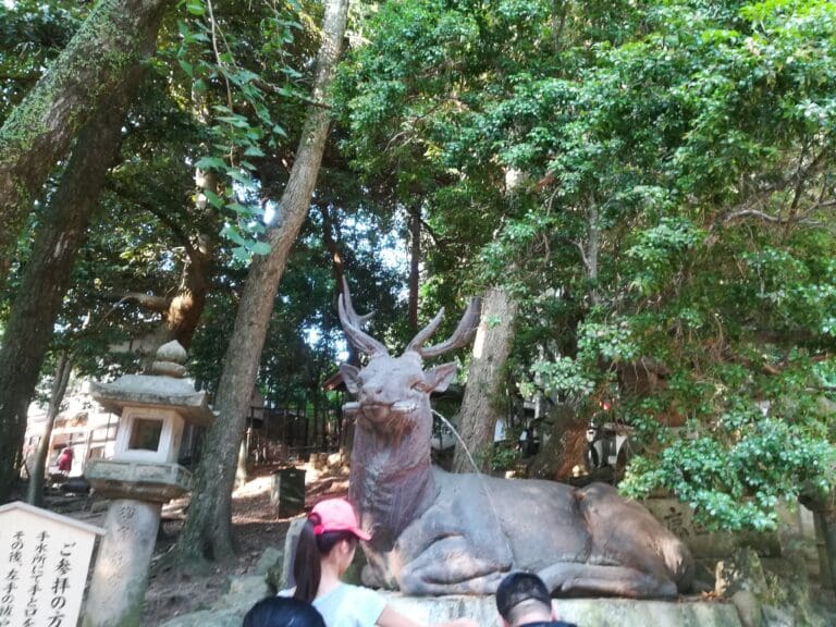 dav Bronze statue of a deer resting among trees at Kasuga Taisha in Nara, Japan, with visitors nearby.