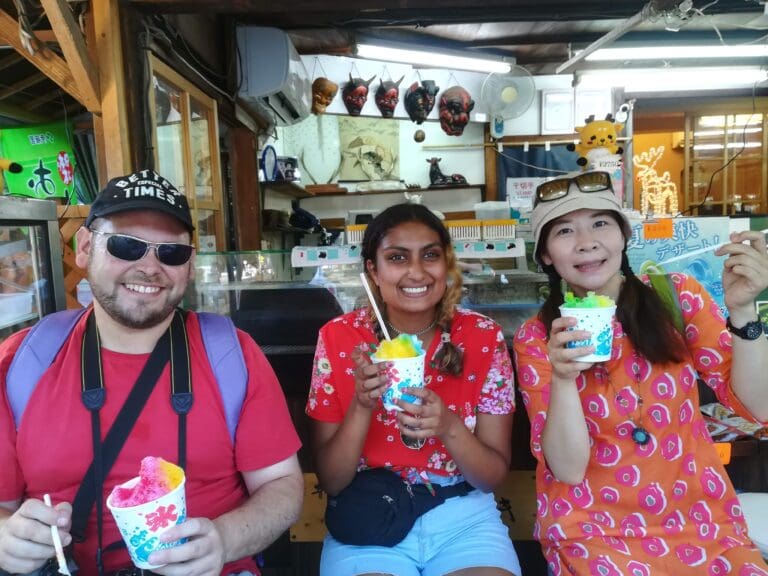 sdr-2 Three individuals enjoying colorful shaved ice desserts while seated at a vendor stall, smiling and holding their treats.