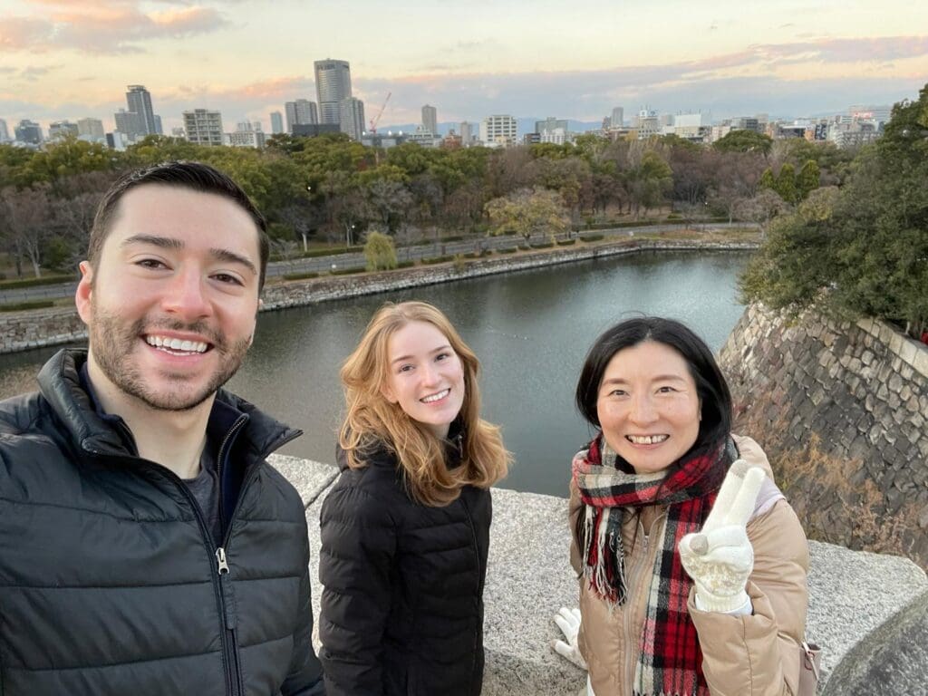 Three individuals posing for a selfie at a scenic location near a body of water, with a city skyline in the background.